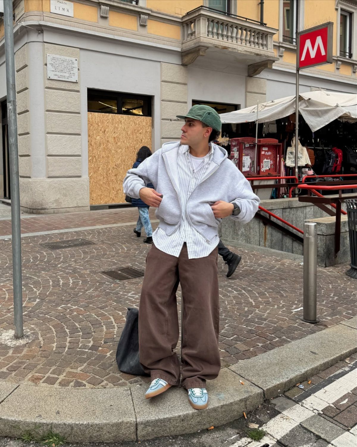 A man wearing an oversized grey hoodie, striped shirt, and wide brown trousers standing on a street.
