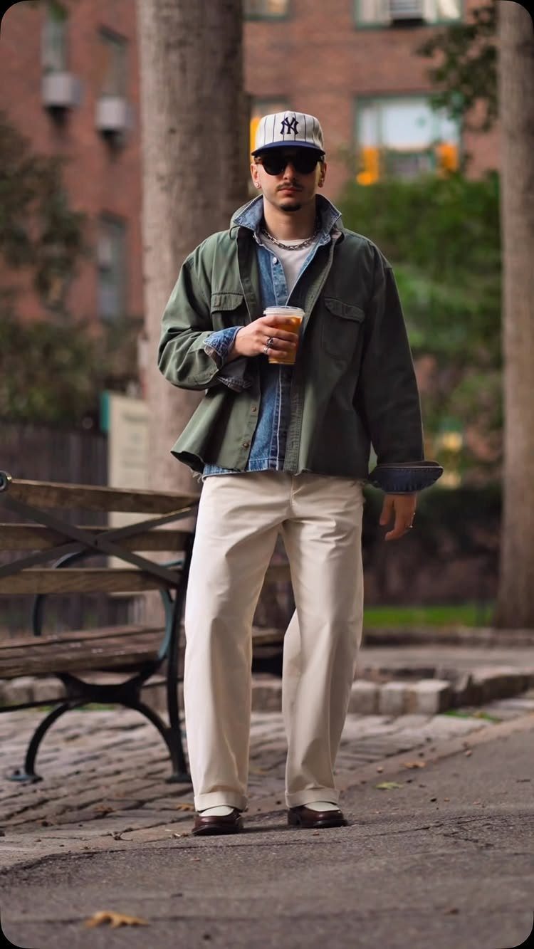 Man wearing a green utility jacket, denim shirt, cream trousers, and a baseball cap outdoors.