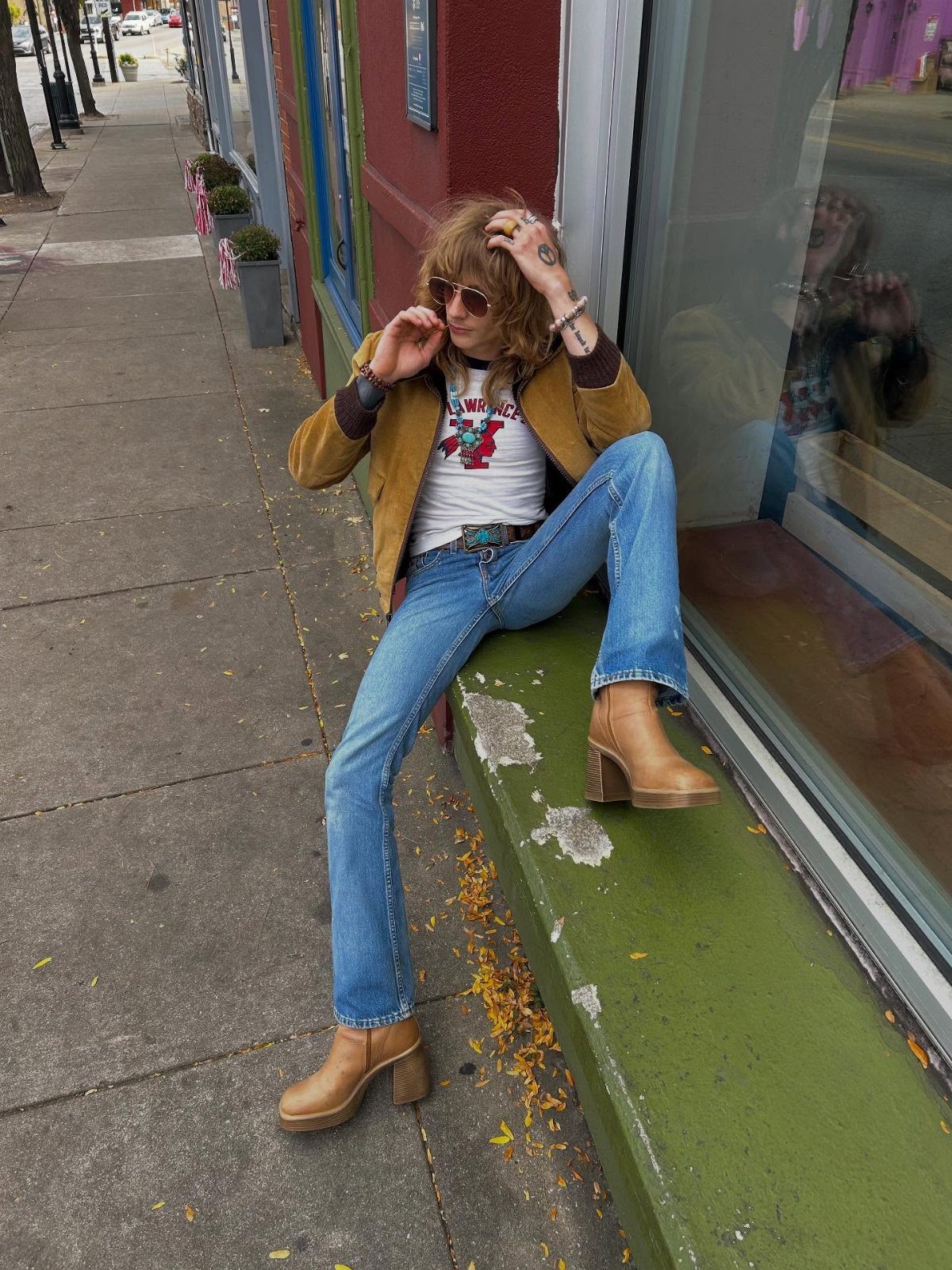 A person wearing a vintage graphic t-shirt, brown jacket, and blue flared jeans sitting on a ledge.