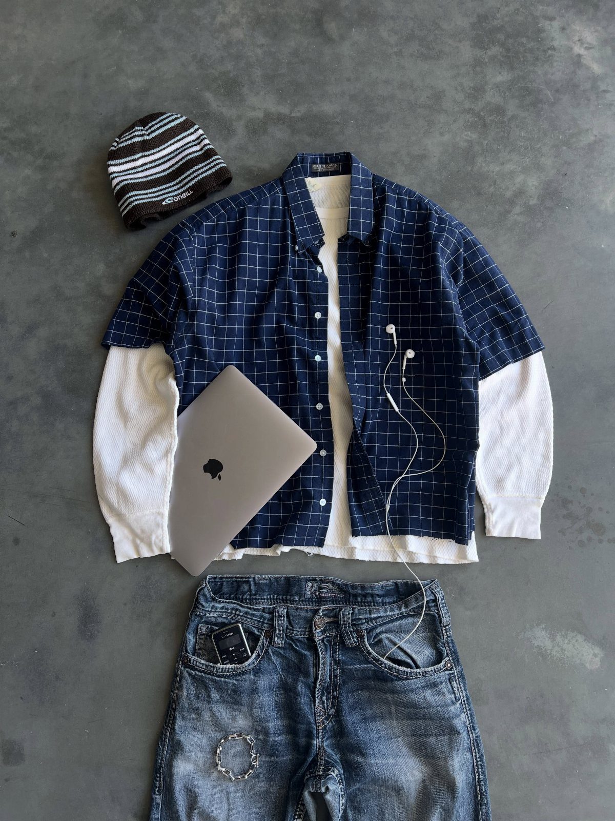 Flat lay of a blue grid shirt, white long-sleeve shirt, distressed jeans, and a striped beanie.