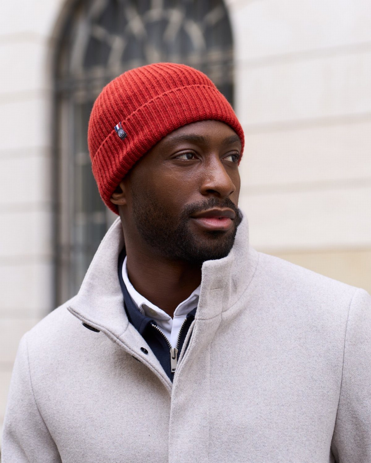 A man wearing a red ribbed knit beanie and a light gray wool coat.