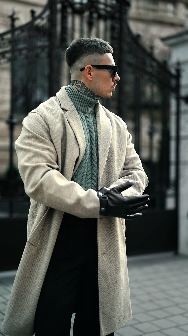 Man in a beige coat, green turtleneck, and black gloves standing outdoors in a city setting.