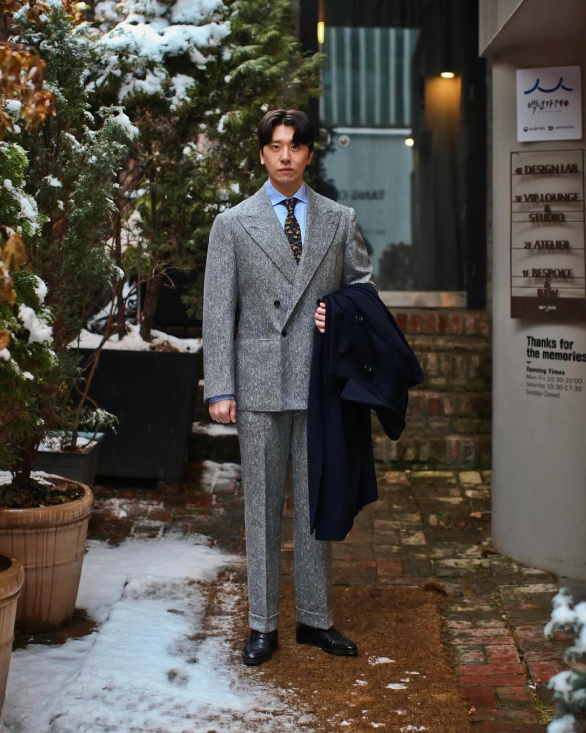 A man posing in a grey tweed suit standing on a snowy brick path.