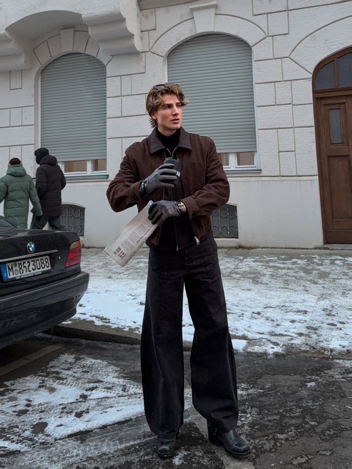 A young man in a brown jacket and black leather gloves holding a newspaper outdoors.
