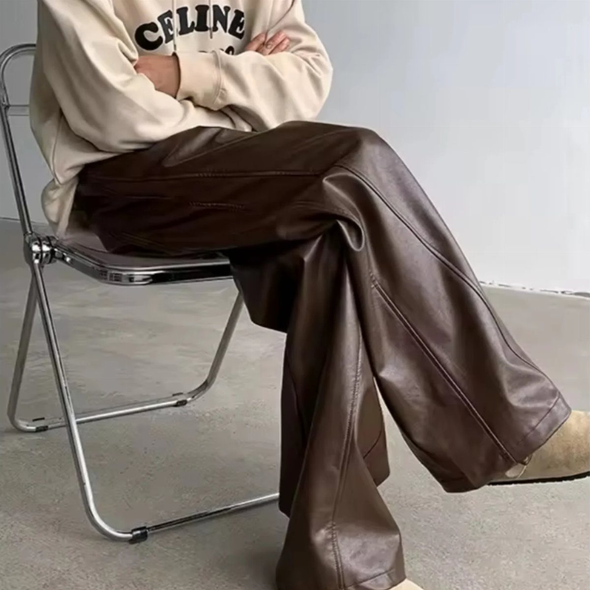 Person wearing a cream Celine sweatshirt and wide-leg brown leather trousers sitting on a chair.