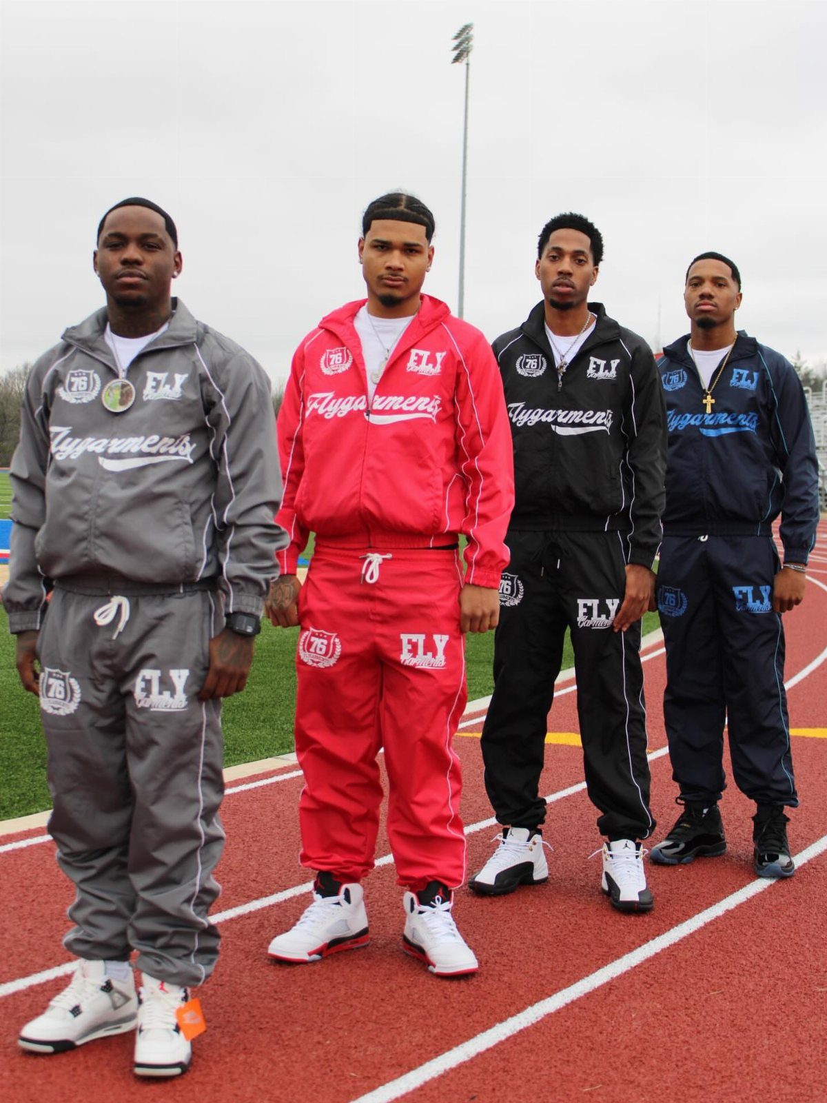 Four men posing on a running track wearing colorful baggy graphic tracksuits and chunky sneakers.