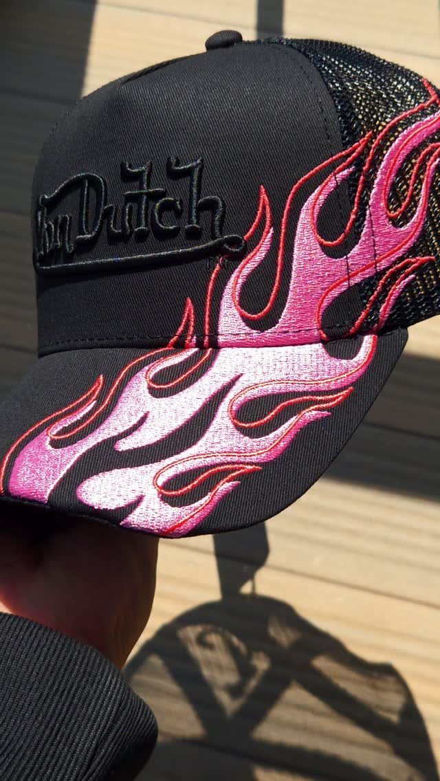 A black Von Dutch trucker hat featuring pink and white flame embroidery held by a hand.