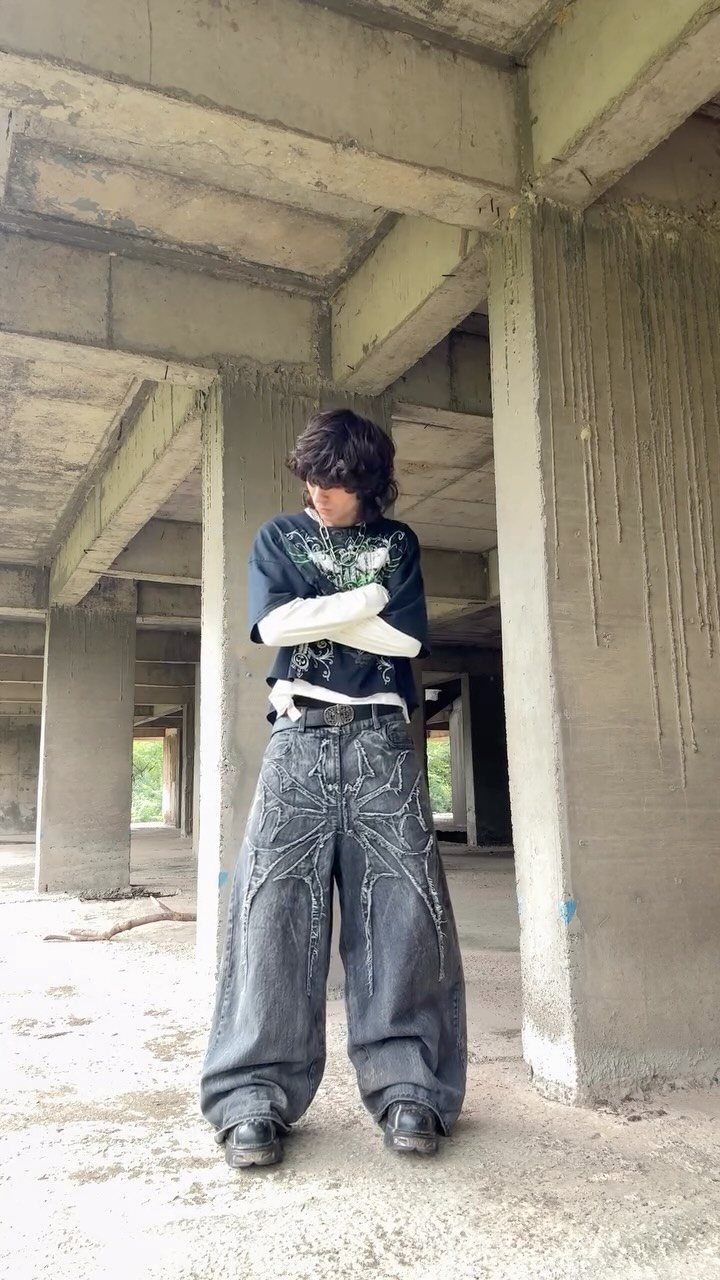 Person wearing oversized graphic streetwear and wide-leg patterned denim pants in a concrete building.