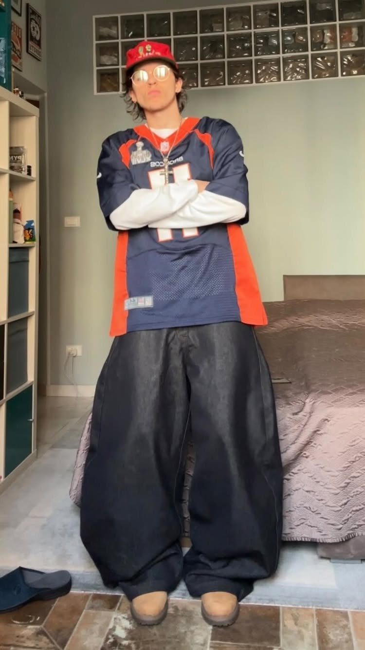 A person posing in a football jersey, extremely baggy black pants, and tinted sunglasses.