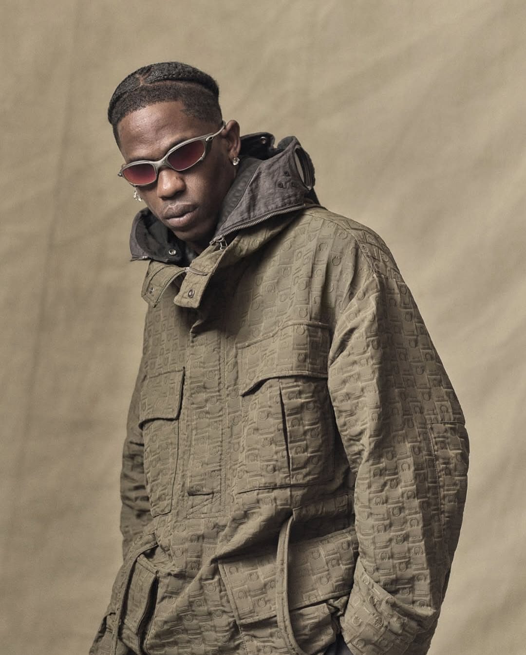 A man in an olive green utility jacket and small sunglasses posing against a tan background.